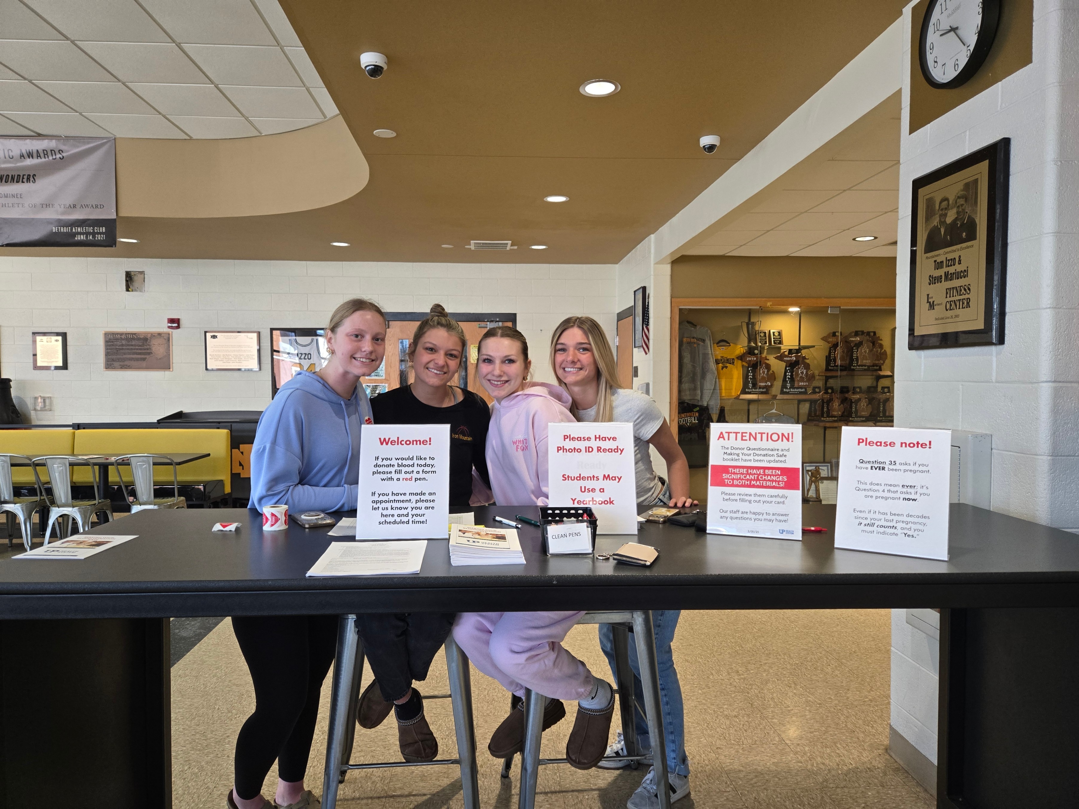 our smiling high school students gather behind a check-in table for a school blood drive. The table displays informational signs regarding blood donation instructions, photo ID requirements, and donor questionnaires.